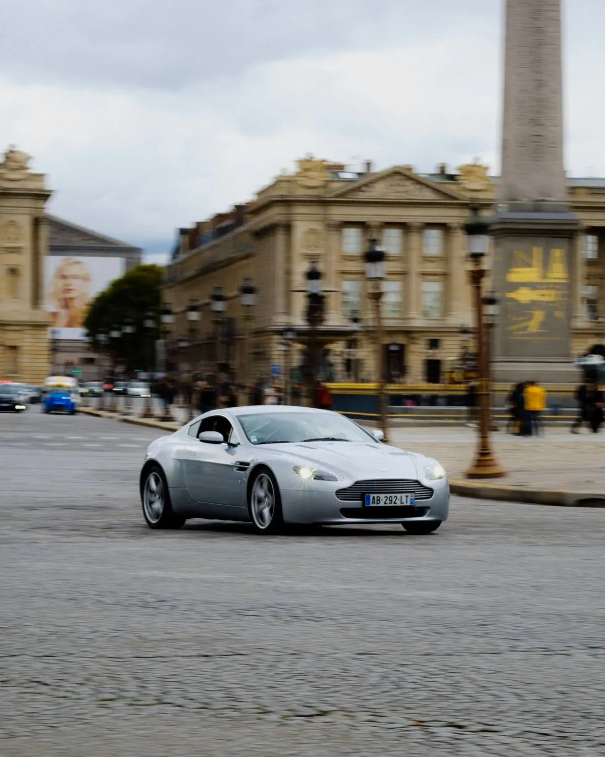 Silver earlier-generation Aston Martin Vantage driving through a city square
